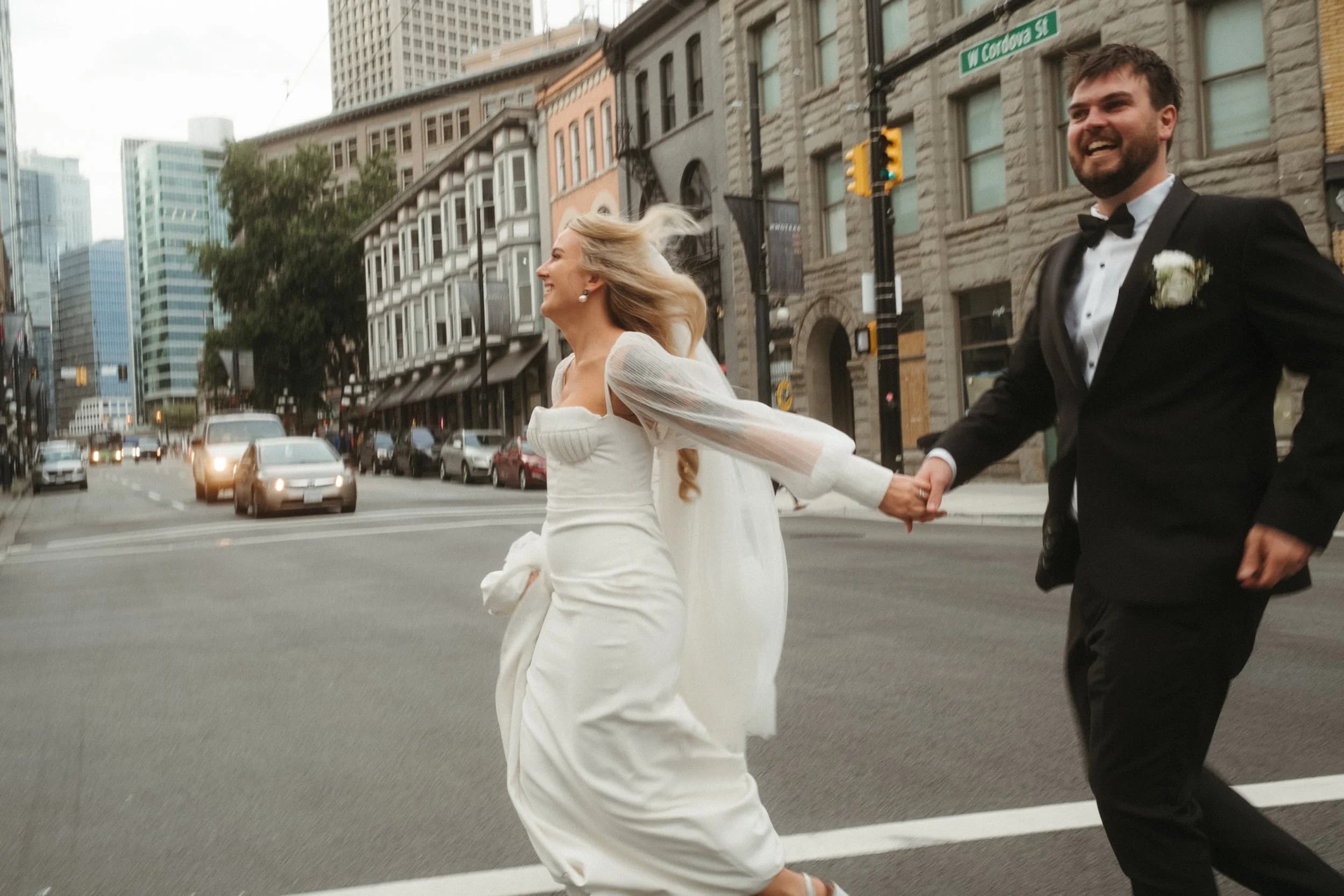 A couple in wedding attire running across a city street.